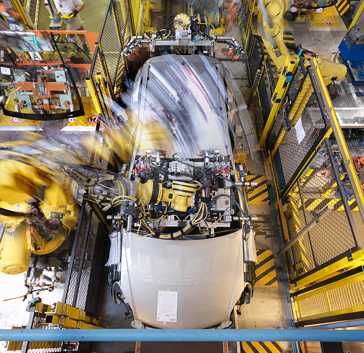 Overhead view of an automobile assembly line featuring a car in the midst of the assembly process. Robotic arms surround the vehicle, actively working on it, while blurred motion indicates rapid activity. The area is enclosed by safety barriers and caution markings for protection.