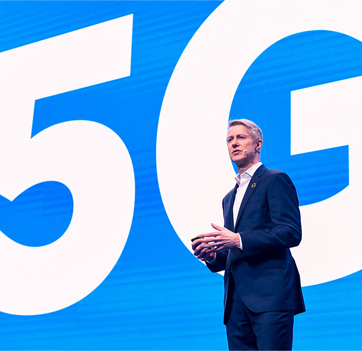 A man in a dark suit gives a presentation on stage in front of a large blue backdrop displaying '5G' in white text. His hands are raised, and he appears to be speaking. The background and his expression suggest a professional or business setting.