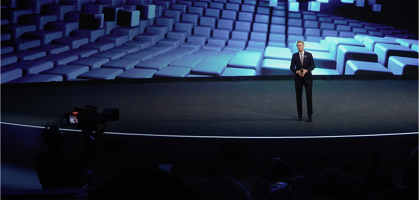 A man in a suit stands on a stage delivering a presentation. The stage is backed by a large, dynamic screen displaying a blue-hued abstract pattern. There is a camera on the left capturing the event. The audience is partially visible in the darkness in the foreground.
