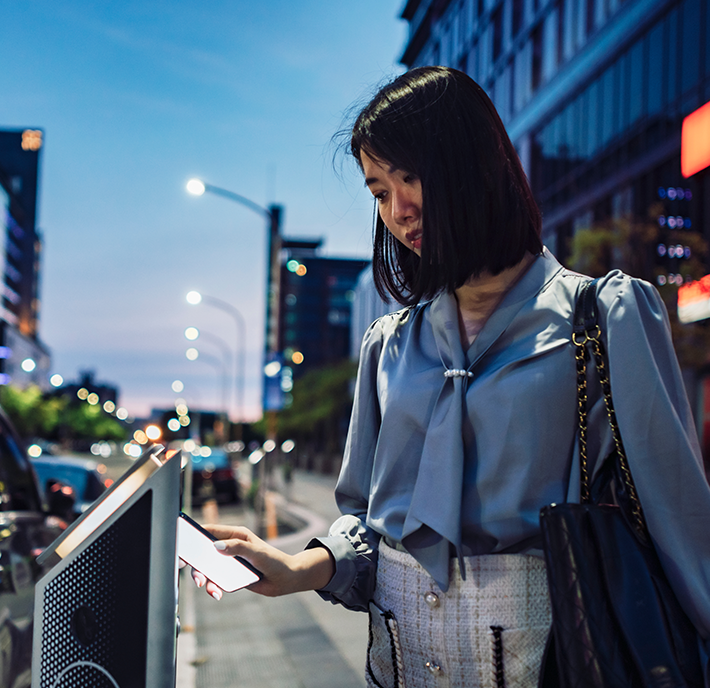 A woman with shoulder-length black hair stands outdoors in an urban area at dusk. She is wearing a light blue blouse and a patterned skirt, holding a smartphone in one hand while interacting with a touch screen kiosk. She has a black bag slung over her shoulder, and city lights illuminate the scene.