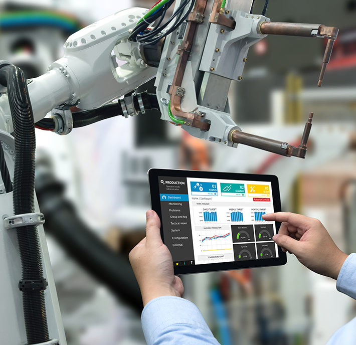 A person is holding a tablet in front of a robotic arm in an industrial setting. The tablet displays various charts and graphs related to manufacturing, such as production performance, machine efficiency, and energy consumption. The robotic arm appears to be stationary, with wires connected to it.
