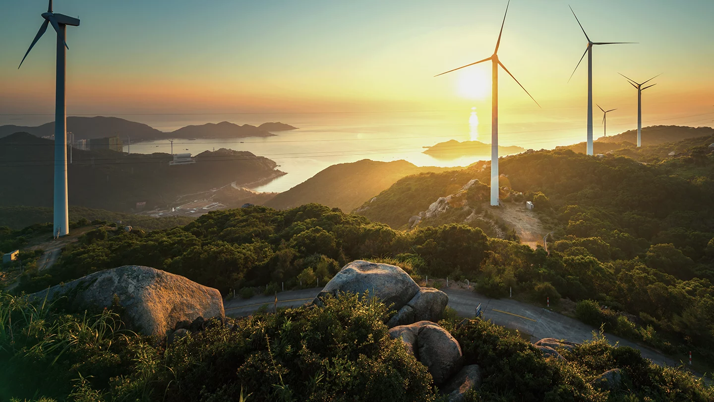A picturesque landscape of a wind farm at sunset, featuring several wind turbines atop green hills. The setting sun casts a golden light over the tranquil ocean and scattered islands in the background. Large boulders and winding paths are visible in the foreground.