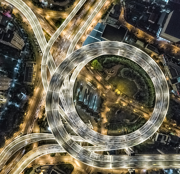 Aerial view of an intricately lit, circular highway interchange at night. Vehicles create light trails on the roads, forming a complex pattern. The interchange encloses a green park with trees, and surrounding city buildings are illuminated, showcasing a dynamic urban landscape.