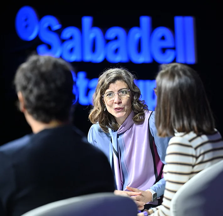 A person with wavy hair and glasses, wearing a purple blouse, speaks to a man and a woman in front of a lit-up "Sabadell" sign. She faces the two people who have their backs to the camera. The setting appears to be a professional or panel discussion event.