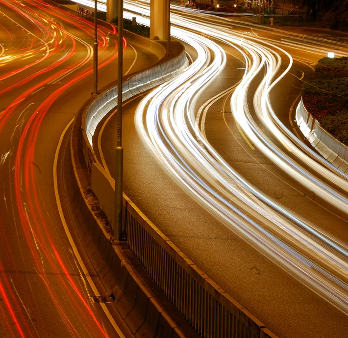 A long-exposure photograph of a busy highway at night, capturing the swirling, colorful streaks of light left by moving vehicles. The lanes curve and converge, with bright white and red trails indicating vehicle headlights and taillights. Streetlights illuminate parts of the road.

.