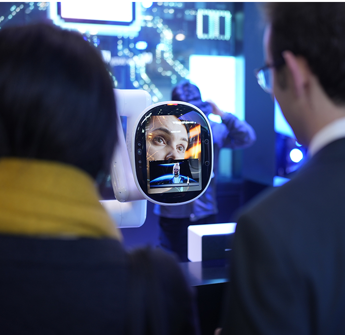 Two people, seen from behind, observe a screen displaying a woman's face and a man in a spacesuit. The setting appears to be a tech or science exhibit with a backdrop of illuminated, futuristic graphics. The woman wears a yellow scarf, and the man wears glasses and a suit.