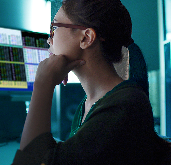 A woman with a ponytail and glasses is working at a computer. She is resting her chin on her hand and looking at a monitor displaying various colorful data charts and graphs. The room is dimly lit with a blue hue. The atmosphere appears focused and analytical.