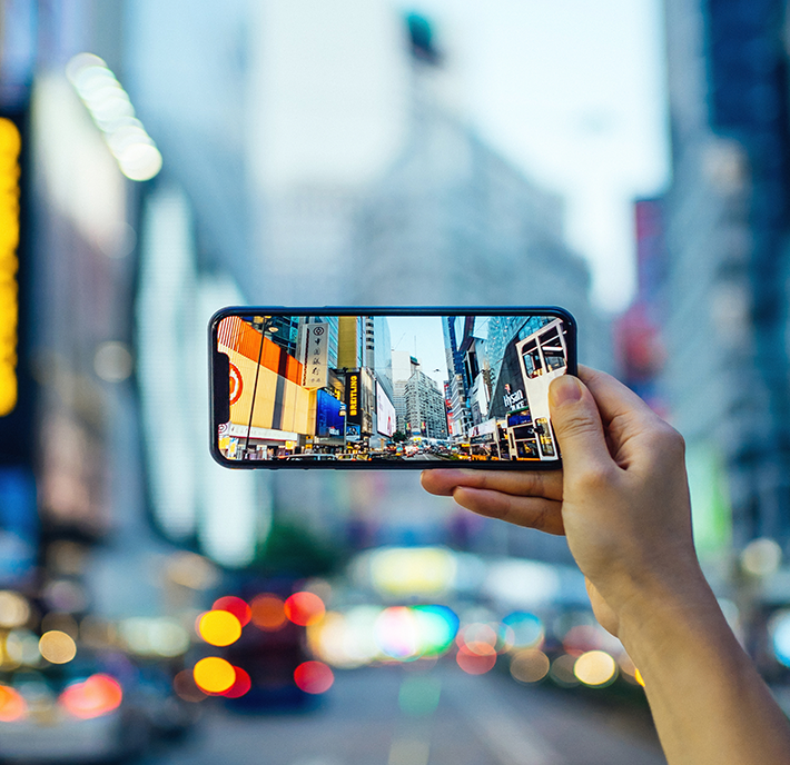 A hand holds up a smartphone, displaying a photo of a bustling, brightly-lit urban scene with billboards and neon lights. The background behind the phone is blurred but appears to be the same cityscape, suggesting that the person is capturing the moment in real-time.