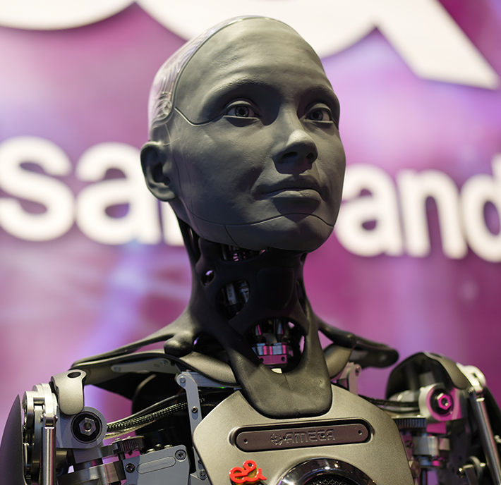 A humanoid robot with a metallic-grey face and exposed mechanical parts stands against a purple background with partially visible white text and logo elements. The robot's realistic facial features include intricately designed eyes, nose, and mouth, giving it a lifelike appearance.