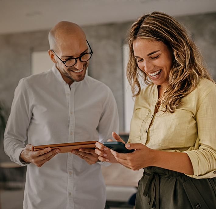 2 people stand together in a modern, well-lit room. onewearing glasses and a white shirt, is holding a tablet. Theother dressed in a light yellow blouse and dark pants, is smiling and looking at a smartphone. Both appear happy and engaged with their devices.