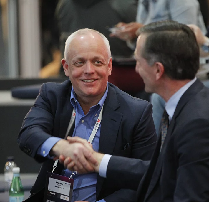 Two men in suits are shaking hands and smiling at each other at a formal event. The man on the left wears a lanyard with an ID badge and a maroon tie, while the man on the right has short dark hair and a patterned tie. There is a water bottle on the table in front of them.