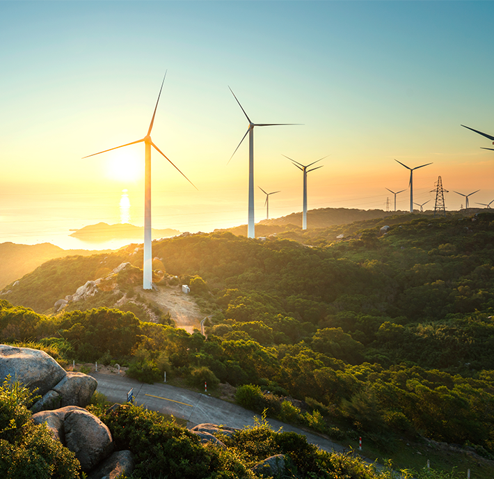 A scenic landscape at sunset featuring several wind turbines on a lush green hill. The sun is setting over a distant body of water, casting a golden light on the surroundings. A mix of trees and rocky terrain is visible in the foreground, with more wind turbines and power lines in the distance.