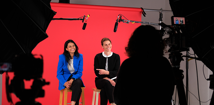 Two women are seated on stools in front of a bright red backdrop, engaged in conversation. One wears a blue blazer and black pants while the other is in a black suit with a white blouse. Professional lighting and recording equipment surround them, with a camera operator positioned in the foreground.