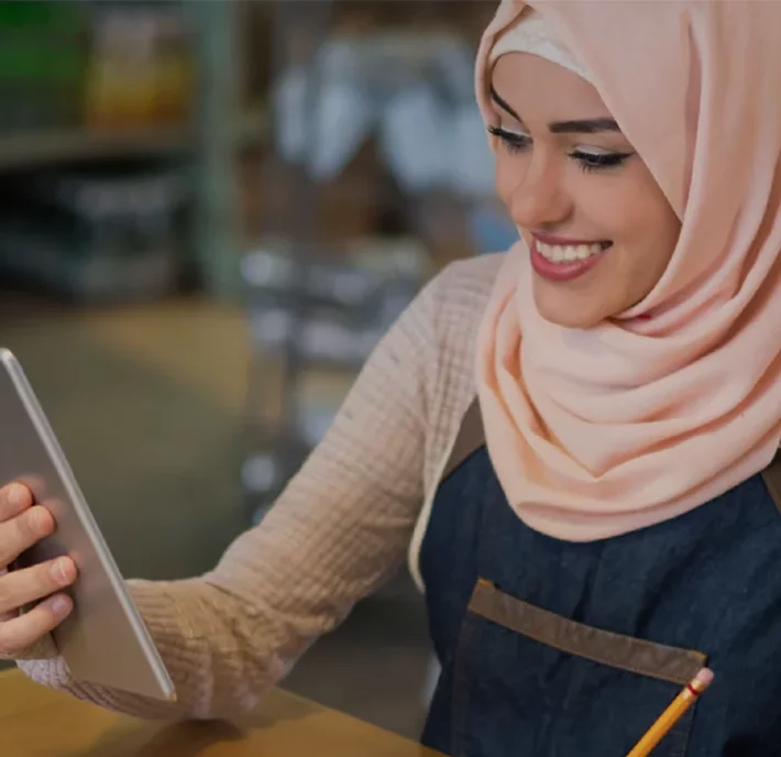 A woman wearing a light pink hijab and a dark apron smiles as she looks at a tablet. She's seated at a table, holding a pencil. The background is softly blurred, suggesting a cozy indoor setting, possibly a cafe or a workspace.