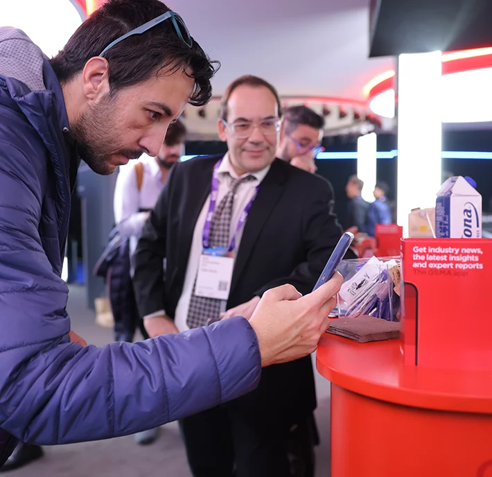A man in a blue jacket leans over a bright red table, engrossed in the GSM App on his smartphone. Another man in a suit and glasses stands smiling in the background. The table features a sign for industry news and packaged items, suggesting an indoor event or conference setting.