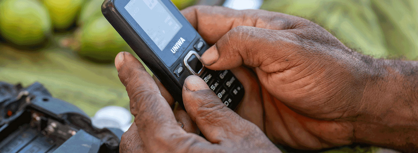 Close-up of a person's hands holding a black mobile phone. The person's fingers are dialing or typing on the keypad. In the background, there are blurred green fruits suggesting an outdoor market setting. The person's skin is textured and weathered.