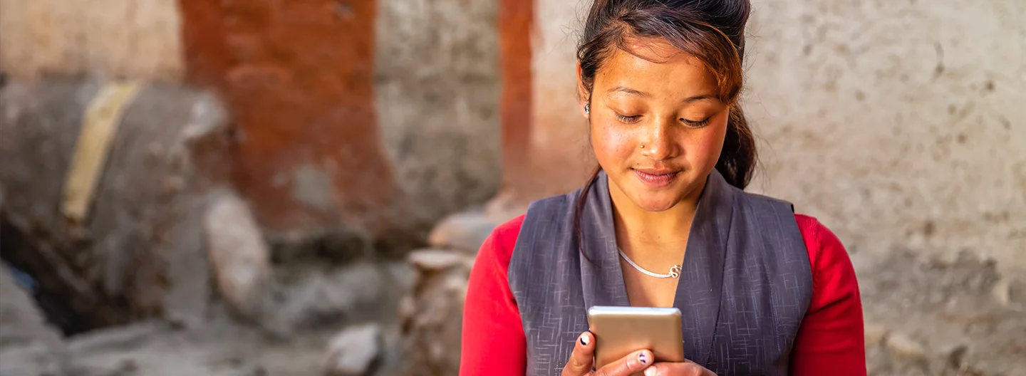 A woman wearing a red top and a gray vest looks at her smartphone with a slight smile. She has long hair tied back, a nose ring, and a necklace. The background features a textured wall in earthy tones, creating a natural setting. The lighting is soft and warm.