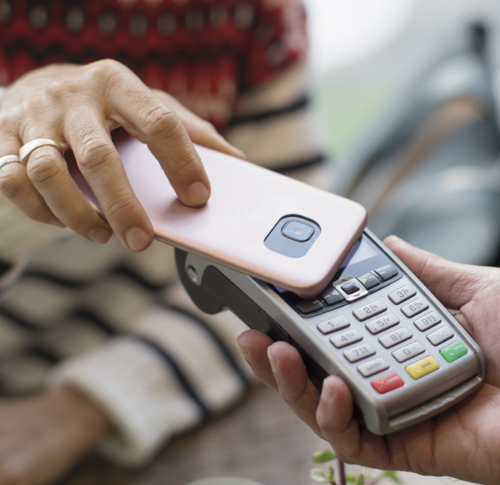 A person uses a smartphone with a pink case for a contactless payment on a card reader held by another person. The card reader has a screen and multicolored buttons. The person holding the phone has a patterned sweater and a ring on their finger.