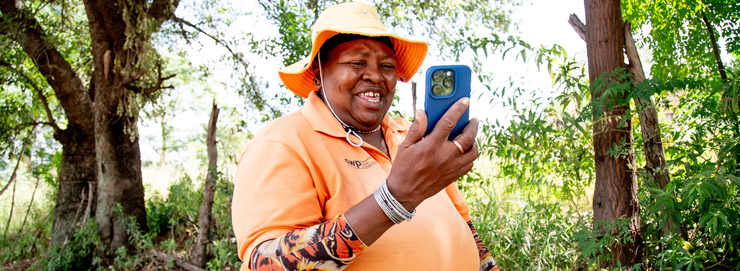 A smiling person wearing an orange hat and shirt stands outdoors among trees, holding a blue smartphone and looking at the screen, with greenery and sunlight in the background.