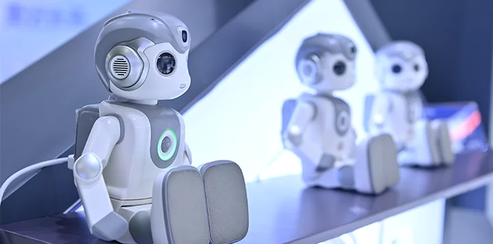 Three small humanoid robots with gray and white bodies sit in a row on a display shelf. Each has a round head, large black eyes, and panel-like legs, with futuristic lighting and a blurred background. The nearest robot is in clear focus.