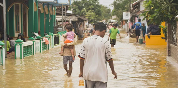 People walk through a flooded street in a residential area. One person carries a child, while others wade through murky water. Houses and fences are partially submerged, and several people stand outside, surveying the situation. Lush green trees line the street.