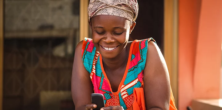 A woman wearing a colorful patterned dress and a headscarf smiles while looking at a mobile phone. She stands in front of an orange wall and a window, appearing happy and engaged with her device.