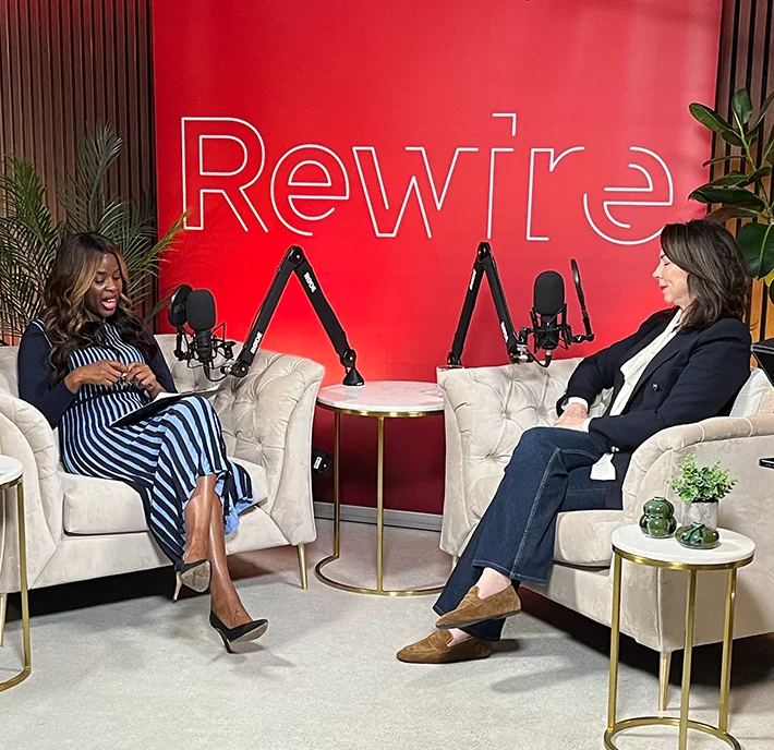 Two women sit in cream armchairs having a conversation in a podcast studio. Both have microphones in front of them. The background is red with the word “Rewire” in large white letters. Green plants and small round tables with decorations are visible around them.