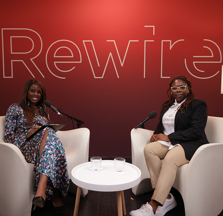 Two women sit facing each other in white chairs, smiling, with microphones in front of them. A small round table with two glasses of water is between them. Behind them, a red wall displays the partial word "Rewire" in large letters.