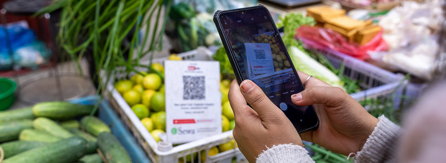A person holds a smartphone and scans a QR code displayed on a sign at a vegetable market stall, surrounded by fresh produce like cucumbers, limes, and leafy greens. The background is filled with colorful vegetables and market items.