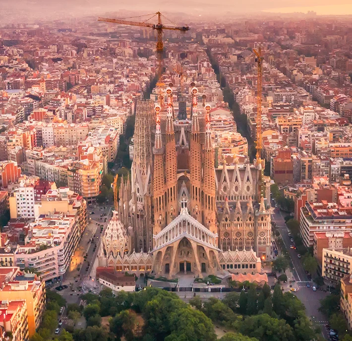 Aerial view of the Sagrada Família basilica in Barcelona at sunset, surrounded by city blocks. The iconic church features intricate spires and ongoing construction with cranes visible. The cityscape extends into the hazy distance under a pink-orange sky.