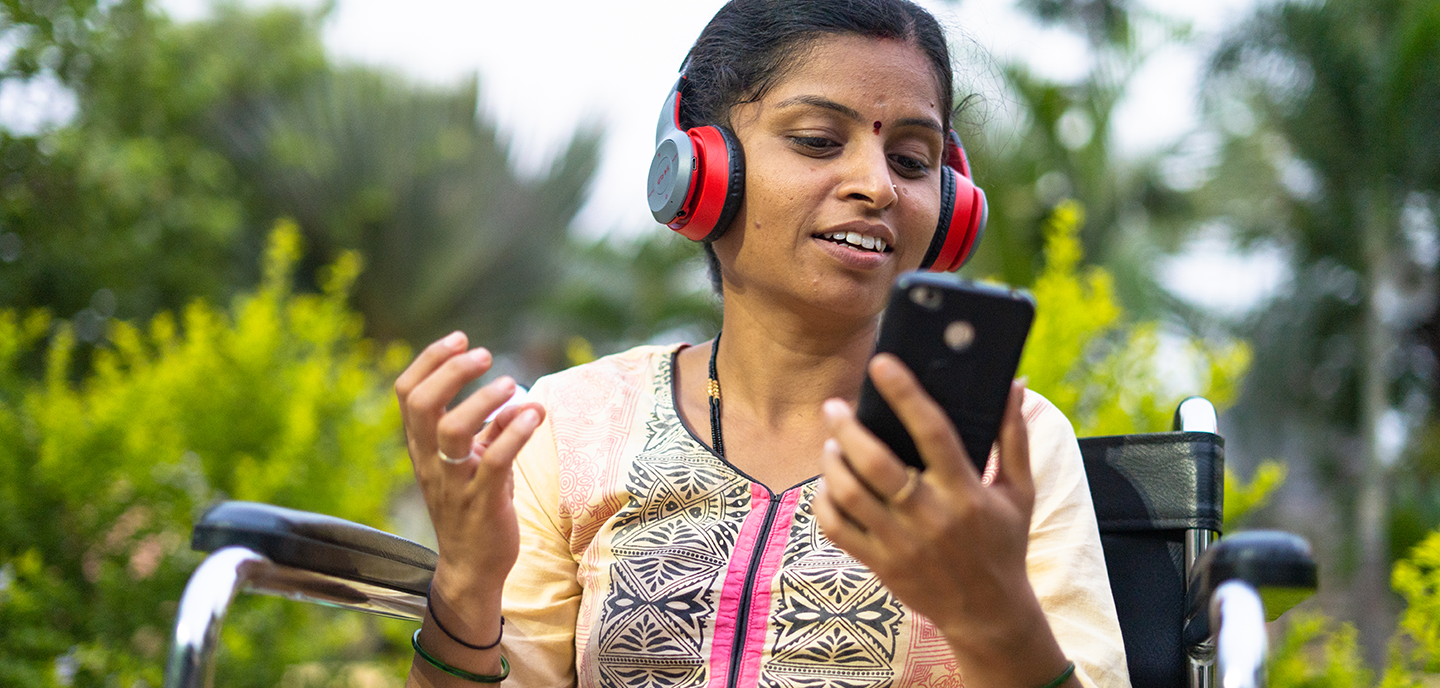 A woman wearing red headphones sits in a wheelchair outdoors, holding a smartphone in one hand and gesturing with the other, smiling and engaged in conversation. Lush green foliage and trees are visible in the background.
