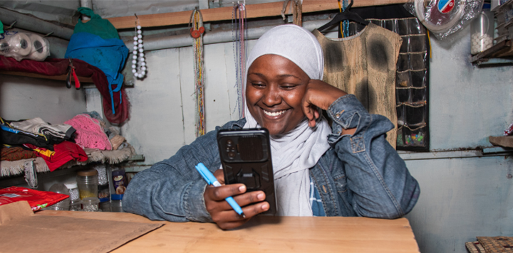 A woman wearing a gray hijab and denim jacket smiles while looking at her smartphone. She holds a blue pen and sits at a wooden table in a small shop with clothes, beads, and household items on shelves behind her.