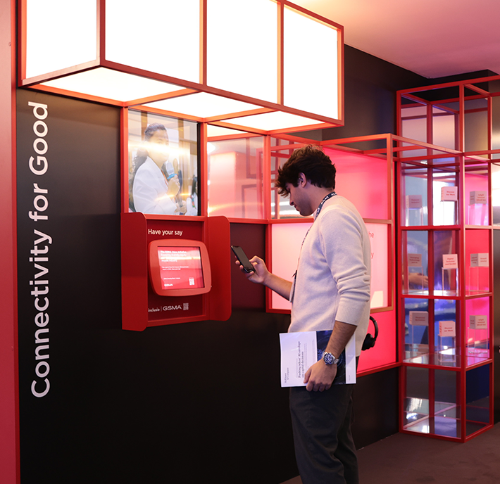 A man in a white sweater stands at a red touchscreen kiosk labeled “Have your say,” using his phone. He holds papers in his other hand. Nearby signage reads “Connectivity for Good.” The scene is set in a modern space with pink and red illuminated decor.