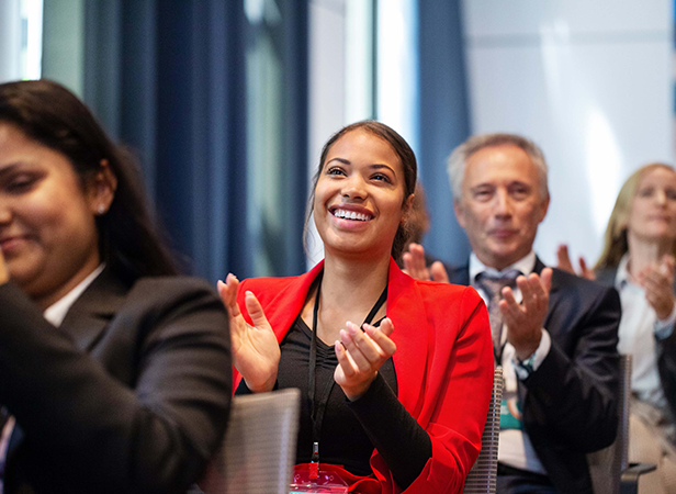 A woman in a bright red blazer smiles and claps enthusiastically while seated at an indoor event. She is surrounded by other people, also applauding, in business attire. The background is softly blurred, suggesting a professional setting.