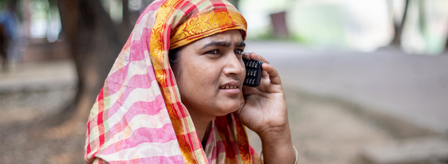 A woman wearing a colourful striped headscarf and patterned clothing talks on a mobile phone outdoors, highlighting the digital divide. She appears focused and concerned, standing in a park area with blurred trees, greenery, and a nearby path.