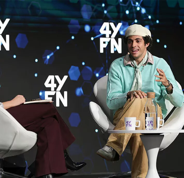 Two people sit in white chairs on a stage with a "4YFN" logo backdrop. The person on the right, wearing a white hat, mint sweater, and khaki pants, gestures while speaking. A carafe and glasses with the "4YFN" logo sit on a small table between them.