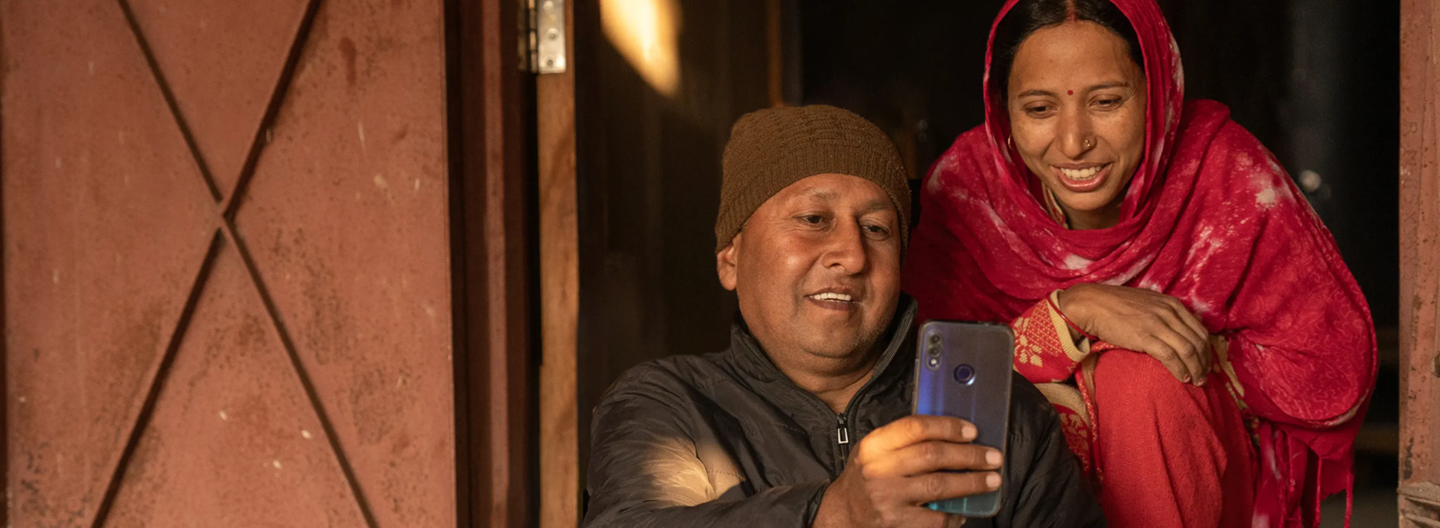 A man wearing a brown beanie and black jacket sits in a doorway, holding a smartphone, while a woman in a red shawl and dress with a bindi leans over his shoulder, both smiling warmly at the phone’s screen. Sunlight softly illuminates their faces.