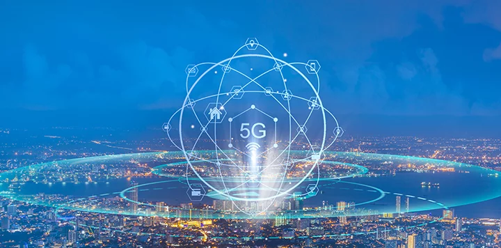 A cityscape at dusk shows illuminated buildings and streets. In the foreground, digital graphics depict “5G” at the center, surrounded by connected icons, symbolizing wireless technology, with glowing blue network rings encircling the city.