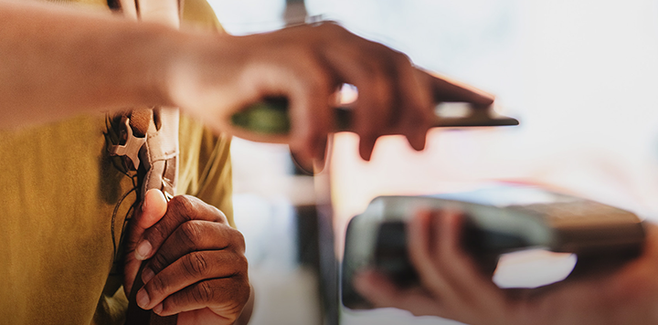 Close-up of a person holding a smartphone near a card reader to make a contactless payment. Another hand holds the payment terminal. The background is softly blurred, focusing on the hands and payment devices. The person wears a yellow shirt and carries a bag.