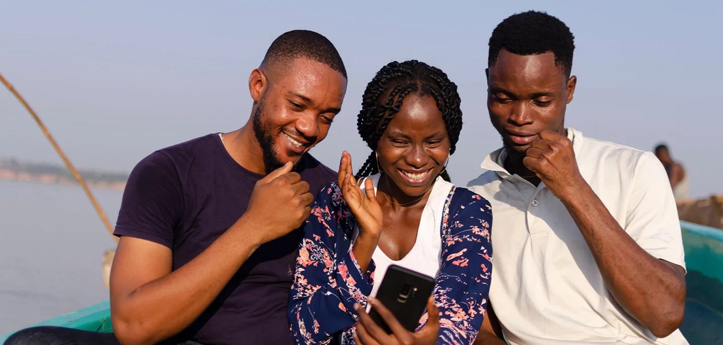 Three people sit closely together outdoors in a boat, smiling and looking at a smartphone held by the woman in the center. The background shows calm water and a clear sky. They appear happy and engaged with what they see on the phone.