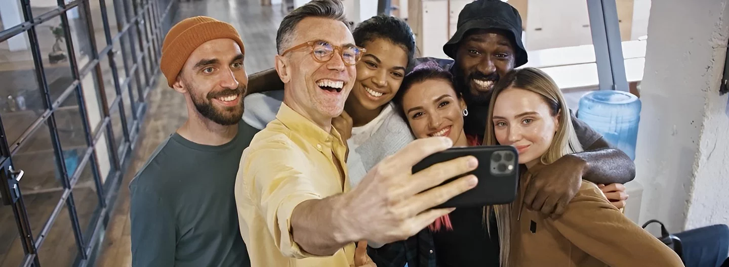 A group of six diverse people stand close together, smiling and posing for a selfie indoors. The person in front holds a smartphone to capture the photo. The setting has large windows and a modern, open-space feel. Everyone looks cheerful and friendly.