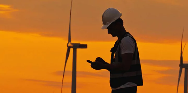 A person in a hard hat and safety vest stands outdoors at sunset, using a mobile device. In the background, wind turbines are visible against an orange sky, suggesting a renewable energy setting.