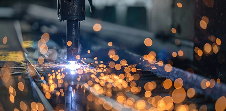 A close-up of a machine cutting metal, producing bright orange and yellow sparks that scatter in all directions. The background is blurred, emphasizing the vivid, glowing sparks and the intense light at the cutting point.