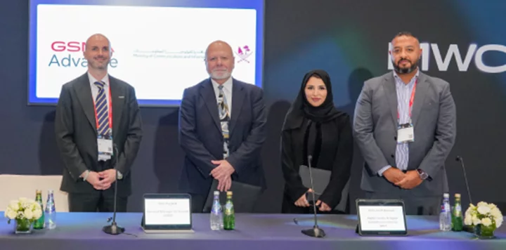 Four people in business attire stand behind a conference table with nameplates, microphones, and bottled water. Behind them, a screen displays logos and text. Two men wear suits and ties, one woman wears a black abaya, and one man wears a gray suit.