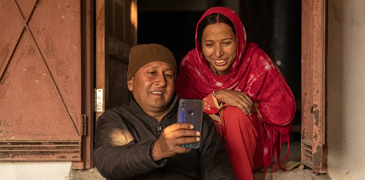 A man in a brown knit hat and black jacket sits by a doorway, holding a smartphone and smiling. Next to him, a woman in a bright red shawl and matching clothes leans in, also smiling as they look at the phone together. Warm sunlight lights their faces.