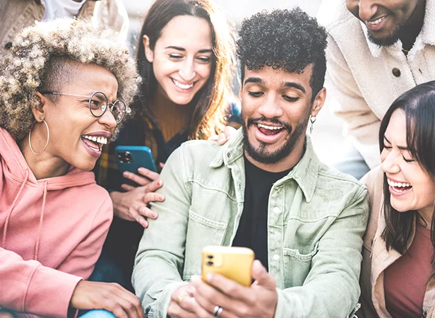 A diverse group of five young adults sit close together outdoors, smiling and laughing as they look at a yellow smartphone held by a man in the center. They appear happy and engaged, enjoying each other's company. Some hold other phones.