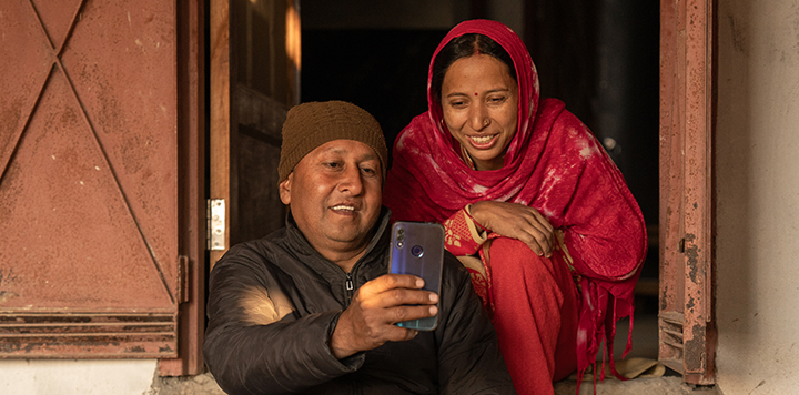 A man in a brown knit hat and dark jacket sits beside a smiling woman in a red headscarf and traditional attire. They sit in a doorway and look at a smartphone the man is holding, appearing to enjoy the moment together. Sunlight filters in warmly.