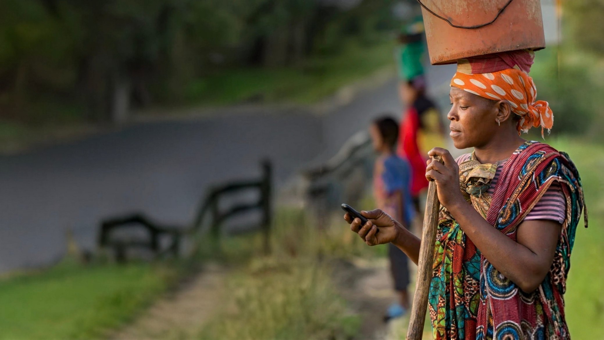 A woman wearing a vibrant patterned dress and orange headscarf balances a container on her head while looking at a cell phone. She stands outdoors near a road, with greenery and blurred figures visible in the background.
