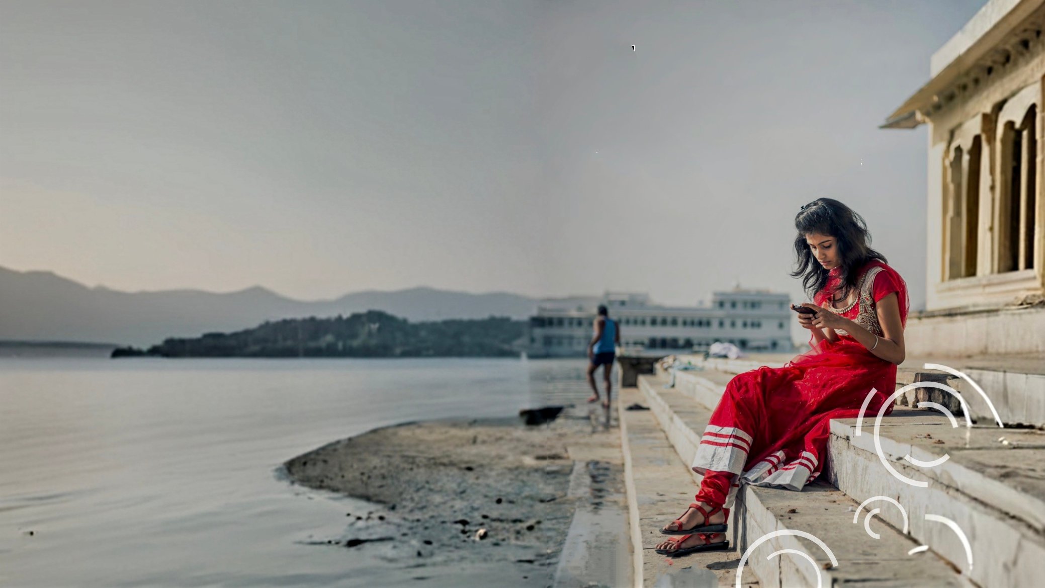 A woman in a red saree sits on stone steps by a calm lakeside, barefoot and looking at her phone. The background shows still water, distant hills, and buildings. The scene feels peaceful and contemplative. White swirl patterns accent the bottom right.