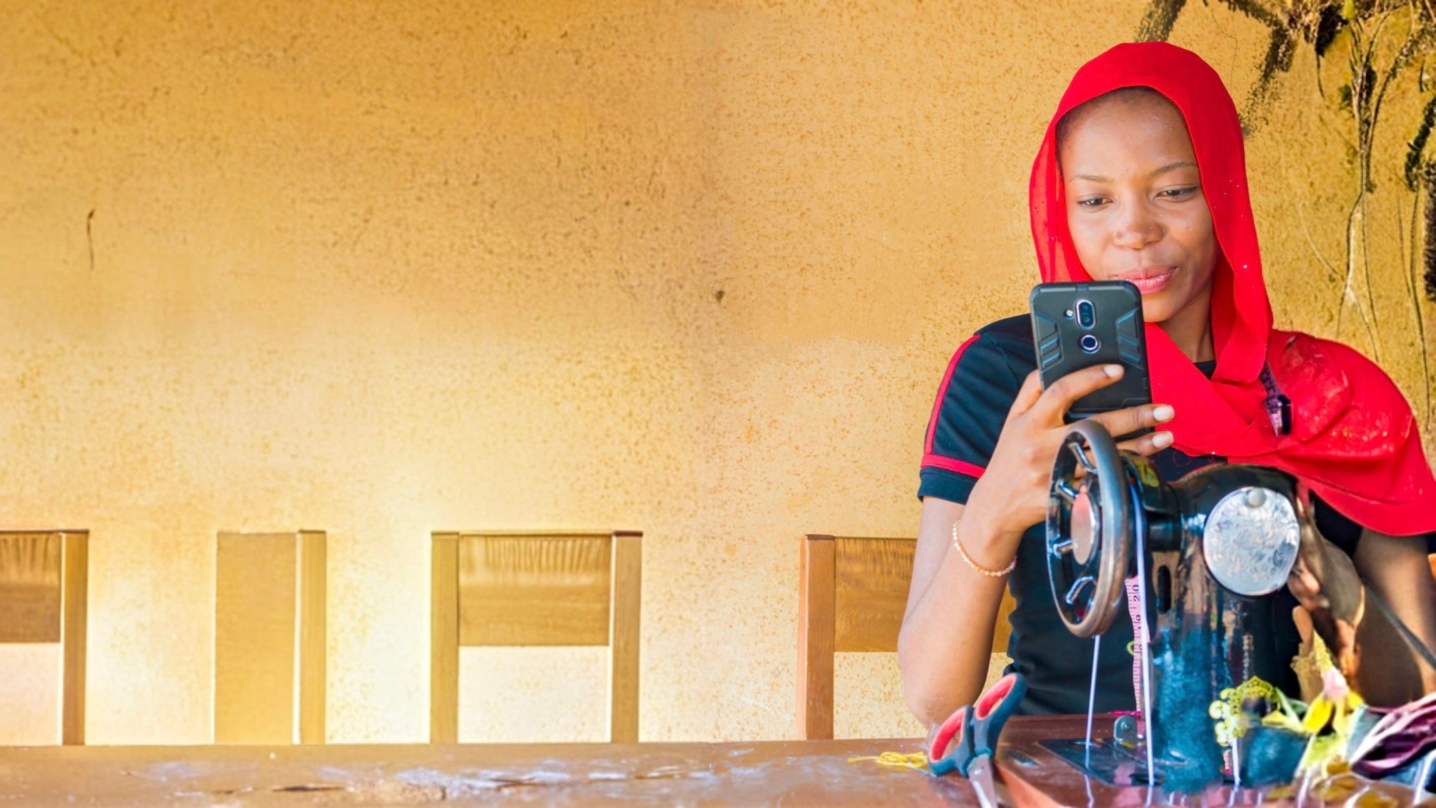 A woman in a red headscarf smiles while using a smartphone, sitting at a sewing machine. She is indoors, with a plain tan wall and wooden chairs in the background. Sunlight casts soft shadows across the scene.